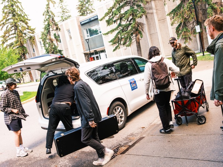 Six people load music and event equipment into a white minivan parked outside a building labeled “University Theatre.” One person lifts a large black case into the van while others stand nearby, carrying bags and a red wagon filled with gear. The group is dressed casually in jackets, flannel shirts, and sneakers, working together under tall evergreen trees on a sunny day. A blue decal on the van reads “Sled Island” suggesting they’re preparing for a festival performance or setup.