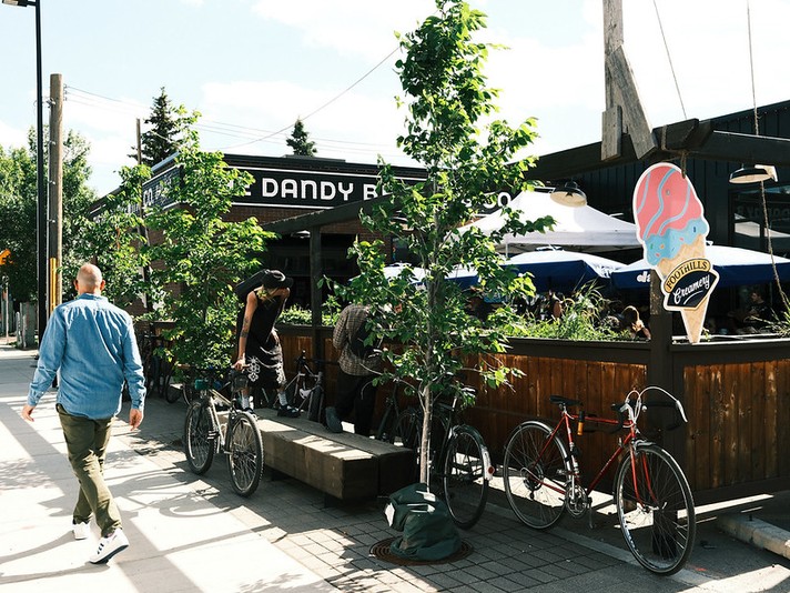 A sunny street scene outside Dandy Brewing Co. during Sled Island 2026 shows people and bicycles along a wooden patio lined with small trees. A person in a blue shirt walks past while another adjusts a bike near the seating area. Several patrons sit under large blue umbrellas enjoying drinks and conversation. A colorful sign shaped like an ice cream cone with pink and blue swirls reads “Foothills Creamery,” adding a playful touch to the lively, relaxed atmosphere.