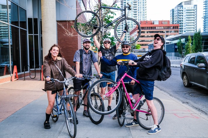 Five people stand on a city sidewalk with their bicycles during Sled Island 2025. One person triumphantly lifts a black Dolan bike overhead, while the others pose with their bikes, including a blue one with a basket and a pink one. Behind them, tall buildings, a parked car, and a tree frame the urban scene, suggesting a celebratory or group ride moment.