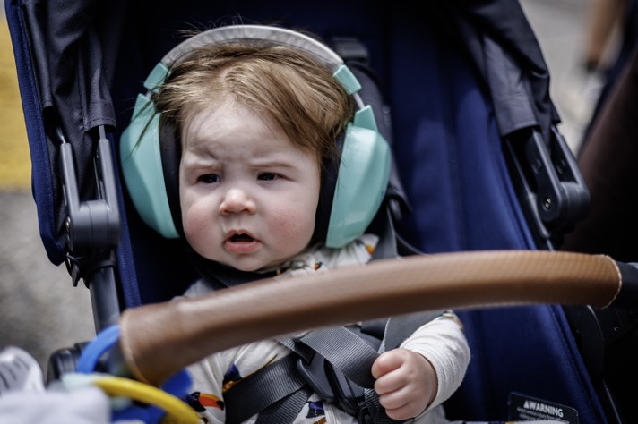 A baby sits in a stroller wearing mint‑green noise‑canceling headphones. The child has light brown hair and rosy cheeks, looking slightly puzzled or curious. They’re dressed in a light patterned outfit and secured with a gray harness. The stroller is dark blue with a brown handlebar in front, and the background is softly blurred, showing an outdoor setting during Sled Island 2026.