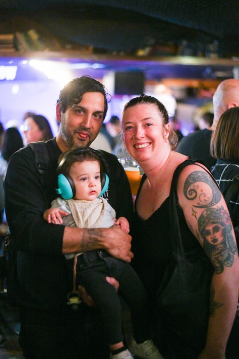 A group of people gathered at Ship & Anchor during Sled Island 2025. One person holds a baby wearing noise-canceling headphones, while another has a visible arm tattoo. The background is softly blurred, with colorful lights and other attendees suggesting a festive, social atmosphere.