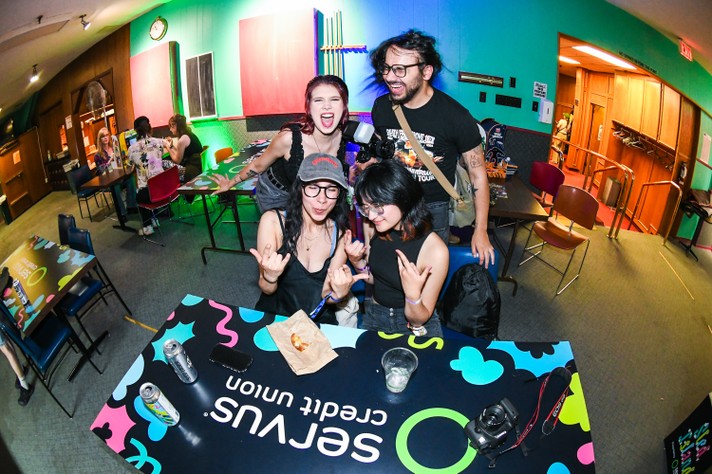 A group of people gather in a brightly colored room with green and blue walls at the #1 Royal Canadian Lehion, decorated for Sled Island Music & Arts Festival. In the foreground, a table displays a “SERVUS Credit Union” design and holds drink cans, a camera, a phone, and a paper bag. Some attendees sit at tables in the background, while others stand and pose for the photo. A clock and various decorations add to the lively, welcoming atmosphere.
