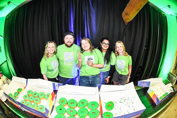 During Sled Isalnd, five smiling people stand behind a table filled with boxes of donuts topped with bright green icing and sprinkles. They wear matching light green shirts that read “Kindness Multiplies,” posing cheerfully in front of a black curtain illuminated by green lighting. The group exudes warmth and positivity, suggesting a community or charity event centered around sharing treats and promoting kindness.