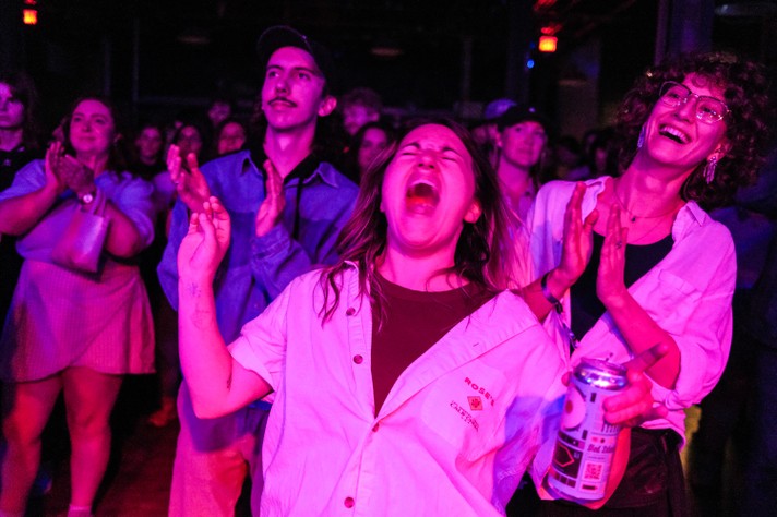 A group of people stand and clap bathed in purple lighting, during a show at Commonwealth for Sled Island 2025. In the foreground, a person wearing a light-colored shirt holds a drink can. The crowd’s expressions and gestures convey excitement and engagement.