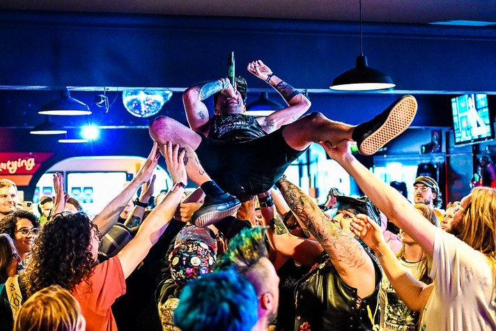 During Sled Island 2026, a performer is crowd surfing in a packed indoor venue, held aloft by dozens of enthusiastic fans. The performer, wearing a sleeveless shirt and shorts, sings into a microphone while gripping it tightly, surrounded by outstretched hands and smiling faces. The crowd’s energy fills the space, illuminated by bright, colorful lights and reflections from a disco ball overhead. The scene captures the raw excitement and communal spirit of live music.