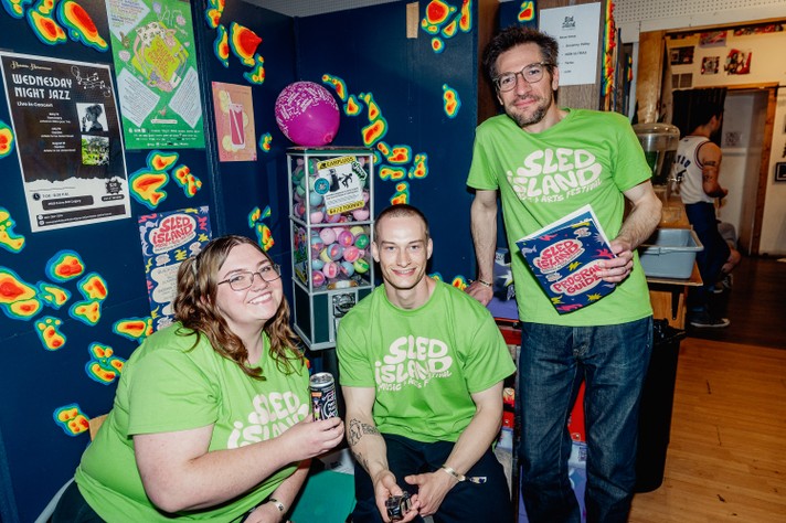 Three people wearing bright green Sled Island Music & Arts Festival T-shirts pose together indoors near a colorful wall covered in posters and heatmap-style decorations. One person sits smiling with a drink can, another kneels holding a small device, and the third stands holding a festival program guide. Behind them, a vending machine filled with pastel capsules and a pink balloon adds playful detail to the lively, creative atmosphere.