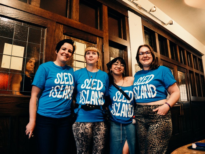 Four volunteers wearing blue “SLED ISLAND” t-shirts stand side by side in front of a wooden wall with glass panels at Central United Church during Sled Island 2025. Each person is dressed casually—one in jeans, one in a skirt, and two in patterned pants. The indoor setting suggests a relaxed moment during the festival.