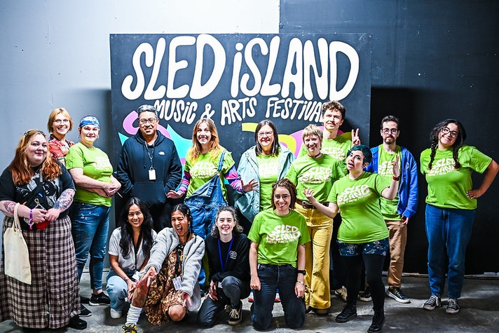 A group of fifteen people pose together indoors in front of a large black sign that reads “SLED ISLAND MUSIC & ARTS FESTIVAL” in bold white letters with colorful shapes behind it. Most of the group wear bright green festival T-shirts, while a few are dressed casually in patterned or denim clothing. They stand and kneel in two rows, smiling and making playful gestures, conveying a cheerful, inclusive team atmosphere.