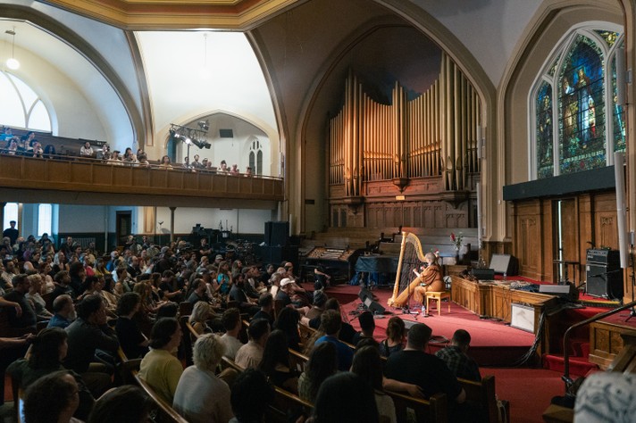 Harpist Mary Lattimore performs during Sled Island 2025 at Central United Church which features high arched ceilings, stained glass windows, and a large pipe organ. The audience, seated in wooden pews and a balcony area, watches attentively. The space is warmly lit, with red carpeting adding to the inviting ambiance.