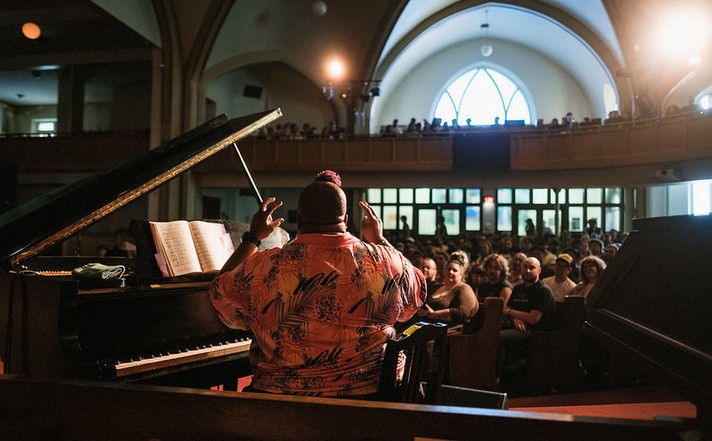 During Sled Island 2026, musician Sharon Udoh performs at a grand piano inside the Central United Church filled with an attentive audience. The performer, seen from behind, wears a vibrant patterned shirt and gestures expressively with both hands as light streams through tall windows behind the crowd. The piano lid is open, revealing sheet music, and the warm lighting highlights the elegant architecture and the sense of connection between artist and audience.