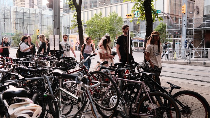 A bustling urban street scene during Sled Island 2025 with people gathered near a bike rack filled with bicycles. Some individuals are chatting, while others walk by. Tall buildings, leafy trees, and a city bus in the background frame the lively atmosphere, highlighting a mix of pedestrian and cyclist activity.