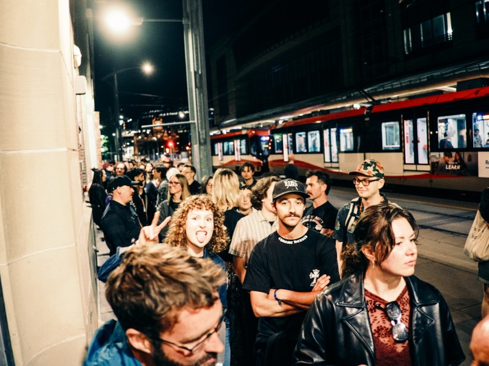A group of people stands on a city sidewalk at night near a tram station outside of the #1 Royal Canadian Legion during Sled Island 2025. The tram glows under streetlights in the background, while individuals in casual clothing wait, chat, and hold bags. The scene captures the energy of a bustling urban evening.