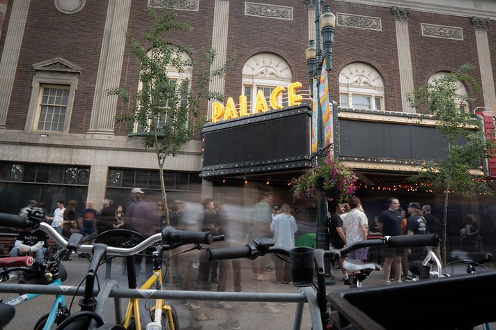 A lively street scene outside the Palace Theatre during Sled Island 2026 shows a crowd gathering beneath its glowing yellow marquee sign that reads “PALACE.” The historic brick building features ornate architectural details and tall windows. Motion blur captures people walking and mingling, adding a sense of energy and movement. In the foreground, several bicycles are parked at a rack, and a lamppost decorated with hanging flowers stands nearby. The atmosphere feels festive and urban, suggesting an event or concert is about to begin.