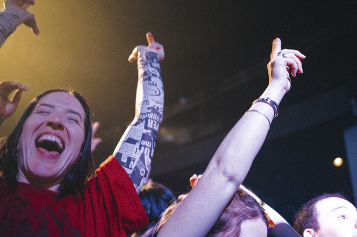 During Sled Island 2026, a person in a red shirt laughs joyfully with their mouth open, surrounded by other concertgoers raising their arms in excitement. One arm in the foreground is covered in a sleeve tattoo featuring bold, newspaper-style text, while another hand points upward toward the stage lights. The warm yellow lighting and blurred background capture the energy and euphoria of a live music performance.
