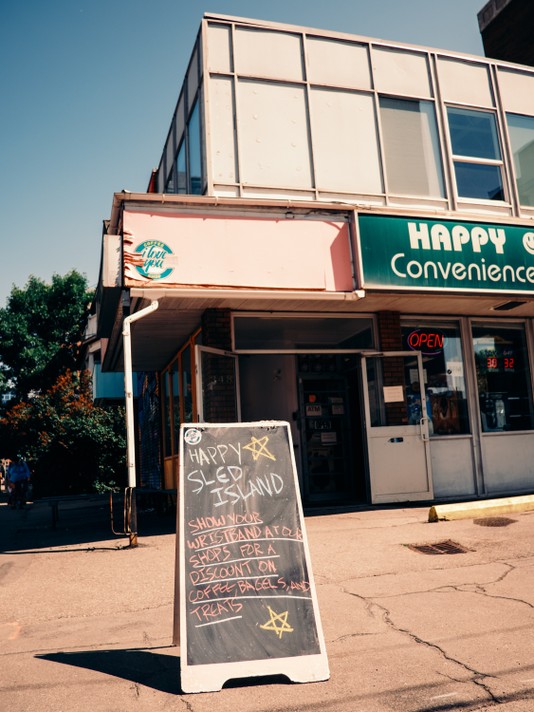 A chalkboard outside of I Love You Coffee Shop reads: “HAPPY SLED ISLAND. SHOW YOUR WRISTBAND AT OUR SHOP FOR A DISCOUNT ON COFFEE, BAGELS, AND TREATS.” In the background to the right, a neon “OPEN” sign glows in the window of a convenience store named “Happy Convenience” with a green sign and white lettering.
