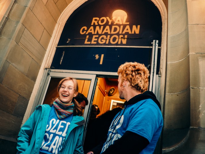 Two volunteers stand outside the entrance of the Royal Canadian Legion building. One wears a blue jacket and a colorful scarf with a blue Sled Island shirt on underneaath, while the other, with blonde hair, wears a blue Sled Island shirt over a black hoodie. Above them, a yellow sign reads “ROYAL CANADIAN LEGION,” marking the venue’s entrance.