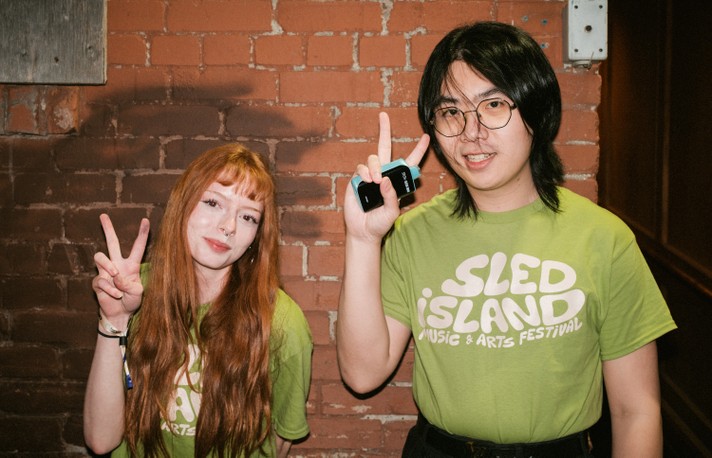 Two people stand in front of a brick wall wearing bright green Sled Island Music & Arts Festival T-shirts. The person on the left has long red hair and a septum piercing, smiling softly while holding up a peace sign. The person on the right has shoulder-length dark hair, glasses, and holds a small device in one hand while raising their index finger. The warm lighting highlights the brick texture behind them, creating a casual and friendly atmosphere that reflects the festival’s creative spirit.
