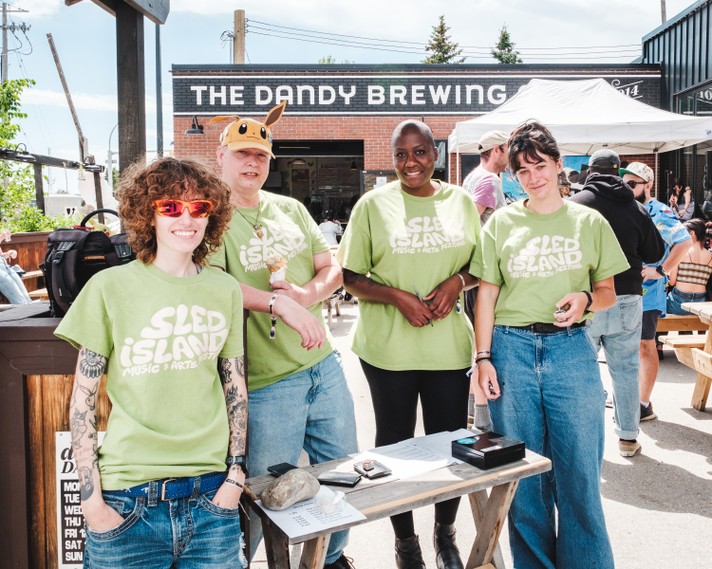 Four people stand outside The Dandy Brewing Company wearing bright green Sled Island Music & Arts Festival T-shirts. They smile and pose near a wooden table with papers and a small cash box, suggesting they’re helping with event check-in or merchandise. One person wears red sunglasses and has tattooed arms, another sports an Eevee hat, while the others stand casually with friendly expressions. Behind them, festivalgoers sit at picnic tables under a white canopy, enjoying the sunny day and lively atmosphere.