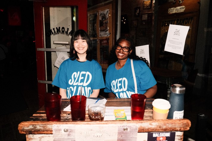During Sled Island 2025, two volunteers wearing blue “SLED ISLAND” t-shirts sit at a wooden table covered with red cups, a glass of dark beverage, papers, and a small box. Behind them, is the entrance to the Palomino, with a red door and a window displaying posters and notices, suggesting an indoor festival.