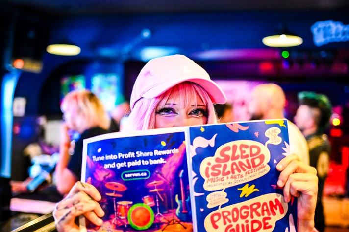 A person with pink hair and a light pink baseball cap holds up a colorful Sled Island Music & Arts Festival program guide inside a lively bar or venue. Their eyes peek playfully over the top of the booklet, which features bright illustrations and bold text. The background glows with neon lights and blurred figures, creating a vibrant, energetic atmosphere that reflects the festival’s creative spirit.