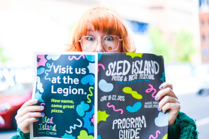 A person with orange hair holds up a program guide for the Sled Island Music & Arts Festival. The guide features vibrant, colorful designs. The left page promotes a venue with the message “Visit us at the Legion. New name, same great pizza,” while the right page displays the festival title, dates (June 19–22, 2025), and presenting sponsor, Servus Credit Union.