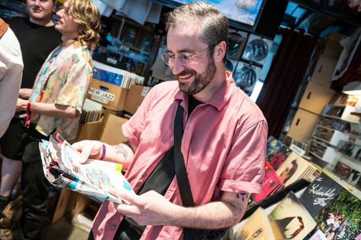 A person in a pink shirt and black shoulder bag browses the Sled Island 2025 program guide inside Sloth Records. Shelves behind them are filled with vinyl records. Another person in a floral shirt browses nearby, adding to the cozy, music-lover vibe of the shop.
