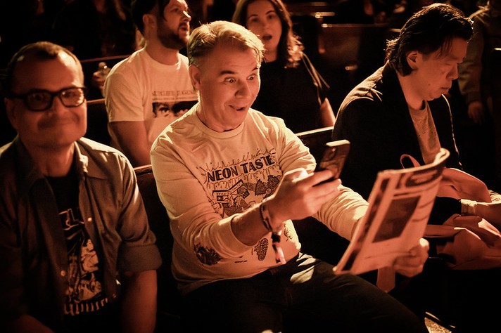 A group of people sit in a warmly lit venue during Sled Island 2026. The person in the center, wearing a white long-sleeve shirt with the words “NEON TASTE” and illustrated graphics, looks at their phone with a wide, surprised expression while holding a printed program or flyer. To their left, a person with glasses smiles subtly, and to their right, another person focuses on something in their hands. The background shows other attendees chatting and watching, bathed in amber light that gives the scene a cozy, energetic atmosphere.