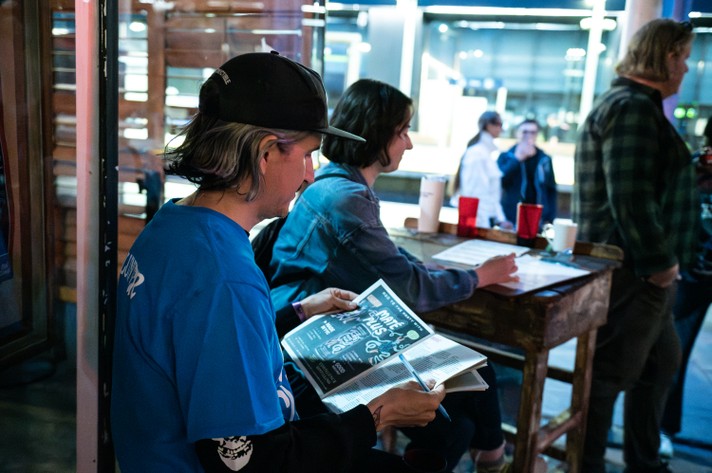 A person in a blue shirt and black cap sits at the front door of the Palomino during Sled Island 2025 and looks through the Sled Island program guide, which features colorful graphics and text. Nearby, another person in a denim jacket writes on paper at a wooden table cluttered with cups, including a red plastic cup and a white one. The background reveals more people and large windows letting in bright light.