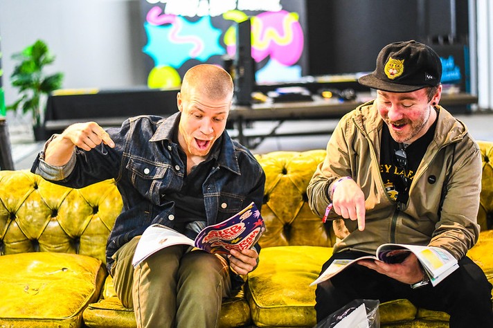 Two people sit on a bright yellow vintage sofa, enthusiastically pointing at pages in their festival program guides. The person on the left, wearing a dark denim jacket, leans forward with an animated expression while holding a Sled Island Music & Arts Festival booklet. The person on the right, dressed in a tan jacket and black cap with a yellow patch, laughs as he gestures toward his own open program. Behind them, colorful abstract artwork and a DJ setup add a lively, creative backdrop to the scene.