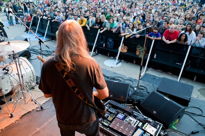 A photo taken from the stage shows J Mascis playing guitar on stage in front of a large crowd at Olympic Plaza during Sled Island 2010. He has long white hair and a brown t-shirt on.