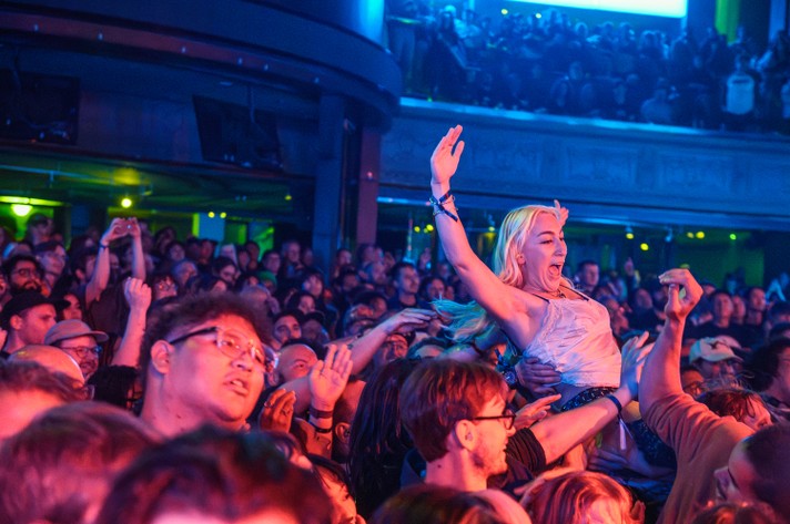 A packed concert at The Palace Theatre buzzing with energy as a person crowd surfs at the Otoboke Beaver show during Sled Island 2025 above a sea of fans. Audience members cheer, raise their hands, and capture the moment with phones. The venue features multiple levels, with spectators filling both the main floor and upper balcony. Colorful lighting enhances the festive, high-energy vibe of the live event.