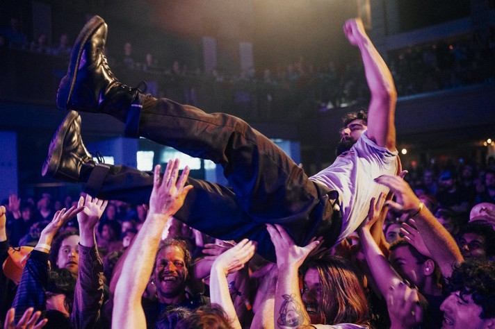 During Sled Island, a person is crowd surfing at a packed concert, lifted high above the audience by dozens of raised hands. They wear black boots, dark pants, and a light shirt, his arm raised triumphantly as they ride the wave of cheering fans. The crowd below is animated and smiling, illuminated by colorful stage lights in purple and gold tones. The background shows a large indoor venue with balconies full of spectators, capturing the electric energy and communal excitement of live music.