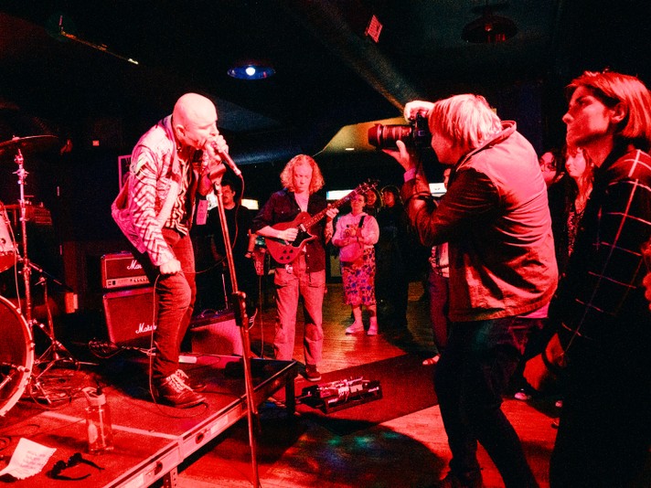 A photo of the band BRIDGELAND performing at in a dimly lit venue called Pin-Bar during Sled Island 2025, featuring a lead singer passionately singing into a microphone while standing on a platform. A guitarist plays in the background, and a photographer with a large camera captures the moment. The audience stands close to the stage, immersed in the show. Red and purple lighting bathes the scene, highlighting musical equipment like a drum set and amplifiers. A water bottle and a piece of paper rest on the platform near the singer, adding to the raw, authentic vibe.