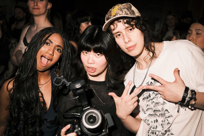 Three friends pose together in a lively indoor crowd at Sled Island 2026. The person on the left, with long braided hair, smiles with their mouth open in a playful expression. The person in the middle holds a professional Canon camera and sticks out their tongue slightly, wearing a black top. The person on the right, wearing a patterned cap, white graphic T‑shirt, and layered bracelets, makes a hand gesture toward the camera. The background shows other people mingling and watching, suggesting a fun, energetic event atmosphere.