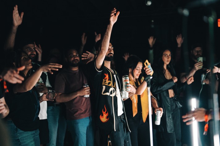 A lively crowd at Commowealth during Sled Island 202t, with people raising their hands and holding drinks. The central figure wears a black shirt with flame designs and the letters “LO,” holding a can and a phone. The atmosphere is energetic and celebratory, with attendees smiling and engaging with one another.