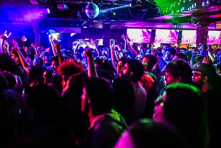 During Sled Island, a packed crowd dances and cheers inside a dimly lit concert venue, illuminated by vivid purple, blue, and green stage lights. People raise their arms and phones toward the stage, capturing the excitement of the performance. A disco ball hangs from the ceiling, reflecting light across the room, while screens in the background display bright visuals. The scene conveys the energy and movement of a live music event in full swing.