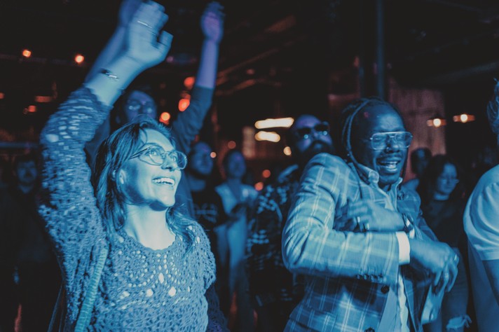 A group of people enjoying a lively concert at Commonwealth during Sled Island 2025, with several individuals raising their hands and dancing. Blue and orange lighting fills the scene, creating a vibrant, energetic atmosphere. The crowd is animated and engaged, with blurred background elements emphasizing motion and excitement.