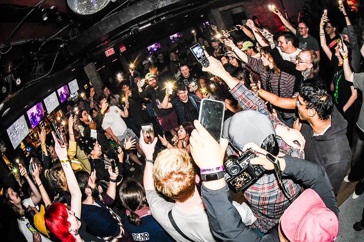 During Sled Island 2026, a lively crowd fills a dimly lit music venue, forming a circle around a central space. Dozens of people raise their phones and drinks high, their screens and flashes creating a sparkling halo of light. The atmosphere is electric and communal, with smiles, laughter, and motion throughout. Multiple TV screens glow in the background, and wristbands show the festival setting. The photo’s wide-angle perspective emphasizes the density and excitement of the moment.