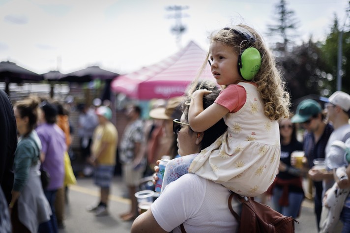 A young child with curly hair sits on an adult’s shoulders at an outdoor event during Sled Island 2026, wearing bright green noise‑canceling earmuffs and a cream dress with a sun pattern over a coral shirt. The adult, wearing sunglasses and a white shirt, holds a drink cup while watching the scene. Around them, people in casual summer clothing mingle near tents and umbrellas under a bright sky, creating a lively festival atmosphere.