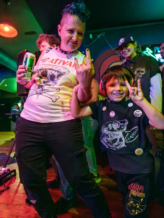 An adult and child pose together at a show at Pin-Bar during Sled Island 2025. The adult wears a white graphic t-shirt with “THE ATIVANS” printed on it, holding a beer can and making a rock-and-roll hand gesture. The child, dressed in a black “HARSH” t-shirt adorned with pins and patches, also makes the same gesture while wearing red headphones. The background is dimly lit with colorful lights and other attendees, suggesting a festive or concert atmosphere.