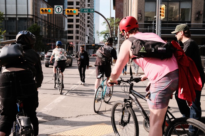 A group of cyclists waits at a traffic light at an urban intersection during Sled Island 2025. They wear helmets, backpacks, and casual clothing. A green arrow on the traffic light signals that they can proceed. The street sign reads “7 Ave SW,” and tall buildings and pedestrians are visible in the background, highlighting a typical moment of city commuting by bike.