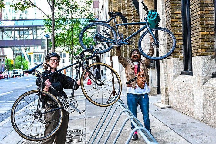 During Sled Island 2026, two people stand on a city sidewalk, each proudly lifting a bicycle into the air near a bike rack. The person on the left wears a helmet, glasses, and dark clothing, smiling as they hold a road bike with thin tires. The person on the right, dressed casually in jeans, a tan jacket, and a T-shirt with a large printed face, raises a blue bike with a teal bag attached to the frame. Behind them are tall brick buildings, trees, and a glass skywalk connecting structures, creating a lively urban backdrop that conveys joy and energy around cycling.