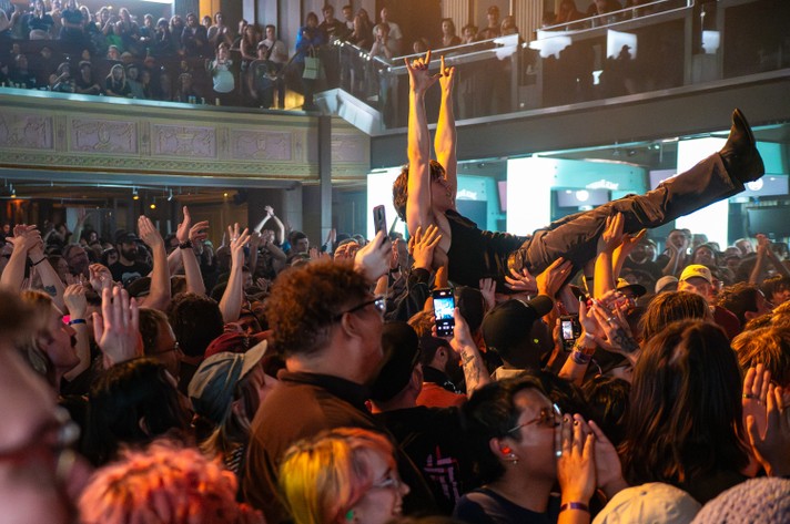 A high-energy indoor concert at The Palace Theatre during Sled Island 2025 with a large crowd cheering and raising their hands. In the foreground, a person crowd surfs above the audience, supported by outstretched arms. Some attendees hold up phones to record the moment. A balcony filled with spectators overlooks the scene, adding to the layered, dynamic atmosphere of the live music event.