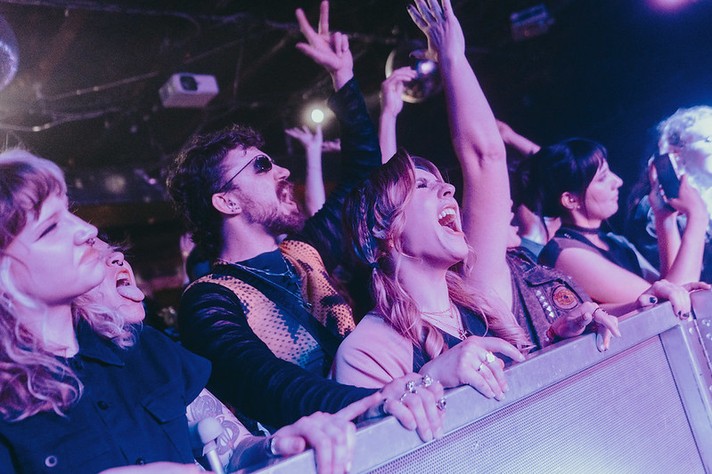 A lively crowd stands pressed against the barricade at a concert during Sled Island 2026, cheering and singing along under purple stage lights. Several people have their arms raised, one flashing peace signs, while others shout joyfully toward the stage. The audience members wear expressive outfits—sunglasses, jewelry, and bold hairstyles—capturing the excitement and energy of live music in an intimate venue.
