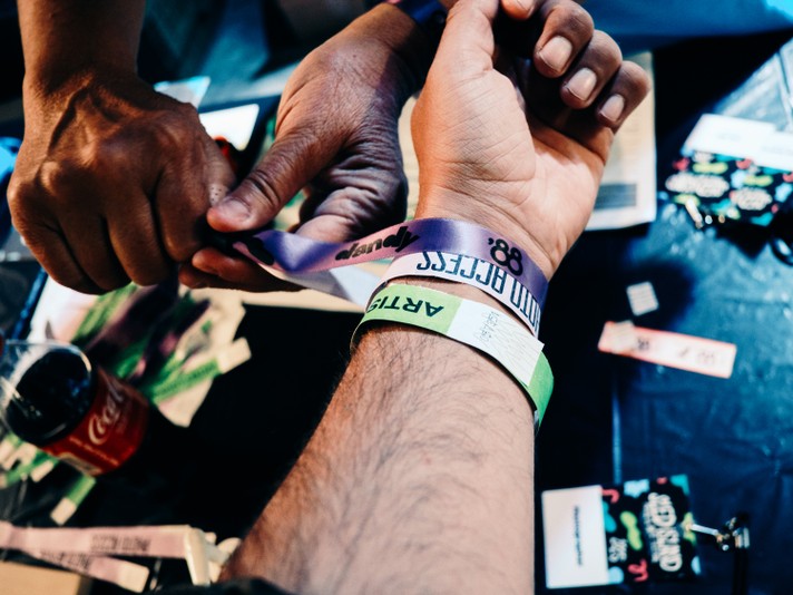 A close-up of a person's wrist being fitted with multiple colorful event wristbands for Sled Island 2025 by another person's hands. The wristbands feature labels like “ARTIST” on a green band and “88 and Dandy” on a purple one. The setting is Sled Island pass pick-up where all attendees check-in and exchange their pass for a wristband. Other wristbands, lanyards, a Coca-Cola can, and event materials are scattered on the surface of the table below the aformentioned hands.