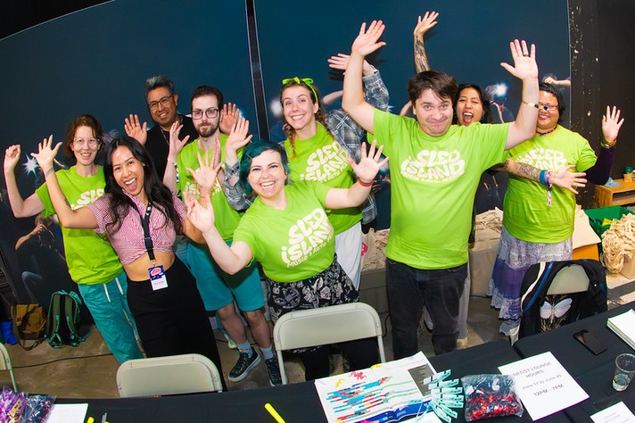 A group of nine smiling people stand behind a table at an indoor event, raising their hands enthusiastically toward the camera. Most wear bright green Sled Island Music & Arts Festival T-shirts, while one person in front wears a pink patterned shirt and a lanyard with an event badge. The table in front of them holds pens, papers, and small items labeled “Artist Check-In.” The lively scene captures a welcoming, energetic team atmosphere at the festival.