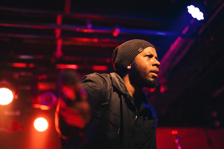 Rapper Arlo Maverick performs at Commonwealth during Sled Island 2025. They are seen wearing a dark jacket and beanie while holding a microphone on stage, illuminated by colorful stage lights. The background includes musical equipment and vibrant lighting, suggesting a dynamic live performance setting.