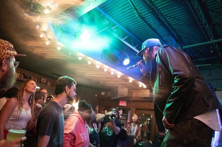 During Sled Island 2026, performer K-Riz is seen wearing a black leather jacket and blue cap leans toward the audience while singing into a microphone at a small indoor venue. Blue and green stage lights beam across the room, creating a smoky, energetic glow. The crowd stands close to the stage, watching intently—some holding drinks, others taking photos. String lights hang from the ceiling, and a red “EXIT” sign glows above a door in the background, adding warmth to the lively scene.