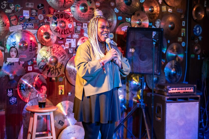 Comedian Tamara Shevon is shown on a stage holding a microphone at Vern's during Sled Island 2025. The wall behind them is decorated with cymbals and a collage of colorful stickers. To the left, a stool holds a glass of dark liquid, and to the right, a speaker and amplifier complete the intimate performance setup.