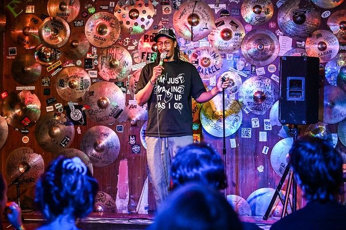 Comedian Faris Hytiaa stands on stage holding a microphone, smiling as they address an audience. The backdrop is covered with dozens of cymbals of various sizes, each decorated with stickers, graffiti, and handwritten messages. The performer wears a black T-shirt that reads “I’M JUST MAKING IT UP AS I GO” and a baseball cap, illuminated by colorful blue and purple stage lights. Audience members are visible in the foreground, watching attentively in the cozy, music-themed venue.